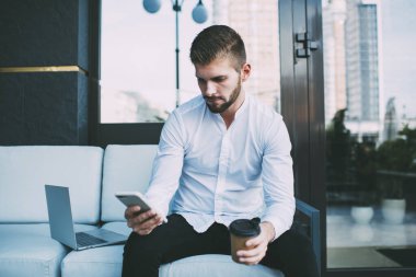 Resmi giysiler içinde, dalgın sakallı bir erkek serbest yazar rahat bir koltukta oturur ve modern kafede kahve içerken akıllı telefonuyla mesajlaşır.