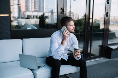 Yoğunlaşmış erkek girişimci, cep telefonuyla konuşurken rahat bir kanepeye oturur ve bir fincan kahve içer.
