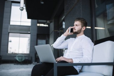 Kendine güvenen sakallı erkek girişimcinin yan görüntüsü kucak reklamında dizüstü bilgisayarla iş sırasında cep telefonundan arama yaparken başka tarafa bakıyor.