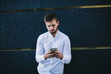 Beyaz tişörtlü ciddi bir yetişkin adam bina duvarına karşı duruyor ve sosyal ağları kontrol etmek için modern cep telefonunu kullanıyor.