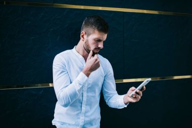 Beyaz tişörtlü, kısa saçlı, konsantre olmamış adam çenesine dokunurken modern cep telefonuna mesaj atıyor ve karanlık duvarın yanında duruyor.