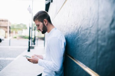 Beyaz tişörtlü ciddi sakallı adamın modern binanın duvarına yaslanırken ve şehir sokaklarında kaldırımda dikilirken akıllı telefon kullanışının yan görüntüsü.