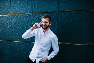 Resmi giyinmiş yakışıklı adam kameraya bakıyor ve gülümsüyor. Akıllı telefondan arıyor ve şehir caddesindeki modern binanın duvarının yanında duruyor.