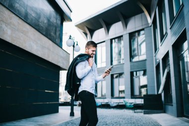Dar bir sokakta, modern binalarla birlikte omuz üstü deri ceketli, cep telefonunu karıştıran mutlu genç bir erkek.
