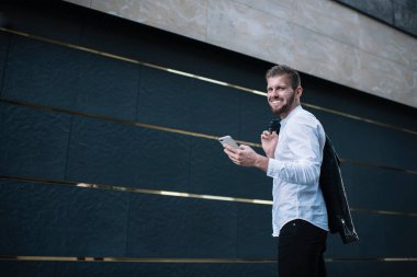 Modayı takip eden sakallı, yakışıklı bir erkek modern ofis binasının yanında dururken cep telefonuyla mesajlaşma takıyor.