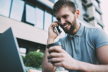 Kafede kahve ve dizüstü bilgisayarla otururken günlük kıyafetlerle konuşan neşeli, sakallı, serbest bir erkek.