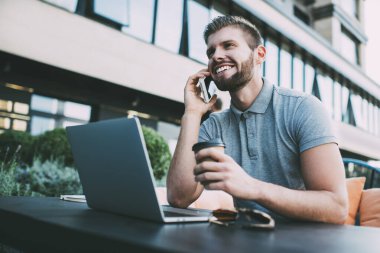 İçerik olarak sakallı, serbest çalışan, günlük kıyafetler giyen, kafede dizüstü bilgisayarla oturan, kahve içen ve telefonla konuşan genç bir erkek.