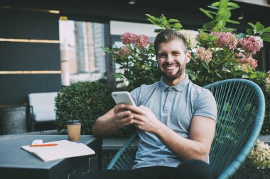 Neşeli, genç, sakallı bir adam. Gündelik kıyafetlerle masada oturmuş cep telefonuyla mesajlaşırken elinde not defteri ve sıcak içecekle.