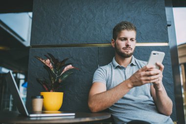 Açık kafede çalışırken laptopla ve saksıyla aynı masada otururken cep telefonuna odaklanmış serbest çalışan bir erkek.