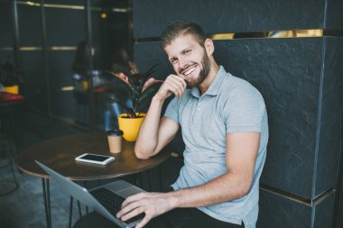 Günlük kıyafetli mutlu bir erkek işçi kameraya bakıyor ve çenesine dokunuyor sokakta masada oturuyor ve dizüstü bilgisayarda yazıyor.