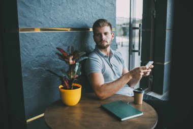 Moda düşkünü, cep telefonu olan genç bir erkek elinde bir fincan kahve ve saksı bitkisiyle masaya bakıyor.