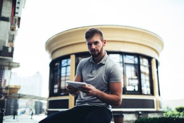 Sokakta sosyal ağları kontrol etmek için tablet kullanan ve bankta otururken kameraya bakan sıradan kıyafetli bir adam.