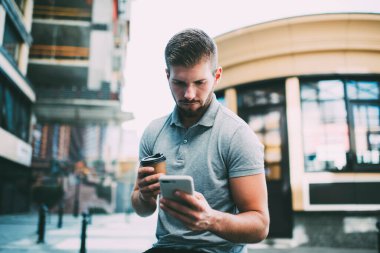 Modern binaların yakınındaki caddede oturan, günlük kıyafetleri içinde kahve fincanı taşıyan, odaklanmış sakallı bir erkek.