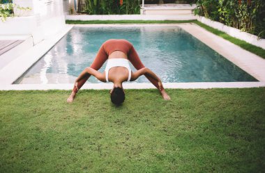 Kız dışarıda poz verirken yoga yapıyor. Uyum ve akıl sağlığı kavramı. Genç ve güzel atletik bir kadın spor giyer ve bahçedeki yüzme havuzunun yanındaki yeşil çimlerde çıplak ayakla dolaşır.