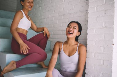 Geniş evdeki merdivenlerde oturan ve dinlenen çok ırklı kadınlara gülmek. Modern kadın arkadaşlığı ve yaşam tarzı kavramı. Spor giyim ve yalınayak güzel Asyalı ve Avrupalı kız arkadaşlar