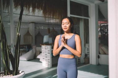 Asyalı kadın yukarıda yoga yapıyor selam duruşu yapıyor ve güneşli evde meditasyon yapıyor. Uyum ve akıl sağlığı kavramı. Spor kıyafetleri giymiş, gözleri kapalı, genç ve güzel bir kızın kısmi görüntüsü.