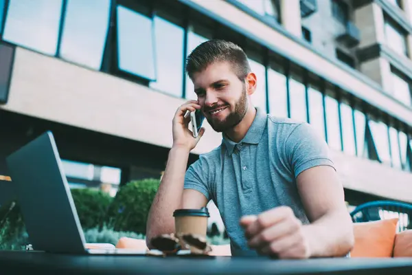 stock image From below of smiling young guy in casual outfit working remotely on netbook and talking on smartphone while sitting on veranda with cup of drink and sunglasses on table