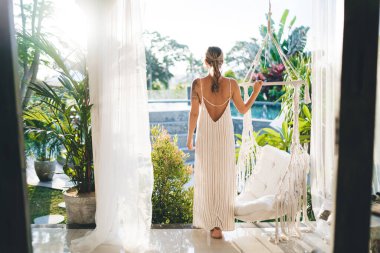 Otelin terasındaki genç bayanın arka görüntüsü. Turizm, tatil ve hafta sonu kavramı. Elbise giyen dövmeli güzel bir kız. Güneşli bir gün. Bali adasında huzurlu ve huzurlu bir yaşam tarzı