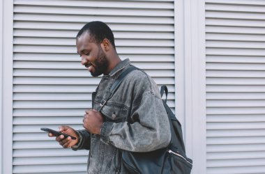 Turistik sırt çantalı mutlu koyu tenli hippi, tatillerde cep telefonu teknolojisini kullanıyor. Neşeli erkek blogcu internet sitesini taramak için 4G 'ye bağlanıyor.
