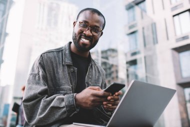 Klasik gözlüklü neşeli erkek programcının portresi dizüstü bilgisayar ve akıllı telefon aygıtı kullanarak uzaktan iş yaparken, cep telefonu ve netbook teknolojisiyle kameraya gülümseyen neşeli bir adam.