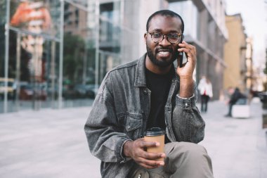 Klasik gözlüklü koyu tenli bir adamın şehir merkezinde hücre sohbeti yaparken kameraya baktığı portresi. Milenyum Afrikalı Amerikalı hippi adam kahve alıp, cep telefonu teknolojisiyle...