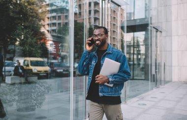 Kapalı dizüstü bilgisayarı olan mutlu siyah göçebe şehirde gezen internet bağlantısını şehir merkezindeki telefon için kullanıyor, gözlüklü, gülümseyen Afrikalı Amerikalı hippi adam
