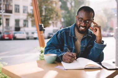 Gülümseyen hippi çocuk not defterinde ödev yazıyor. Kaldırımdaki kafede eğitim zamanının keyfini çıkarıyor. Klasik gözlüklü mutlu Afro-Amerikan erkek öğrenci tasarım projesi için skeç hazırlıyor.