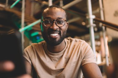 Boş zamanlarında cep telefonu kullanan neşeli hipster adamın portresi. Optik gözlüklü mutlu Afro-Amerikan adam akıllı telefon cihazı tutuyor ve kameraya gülümsüyor.