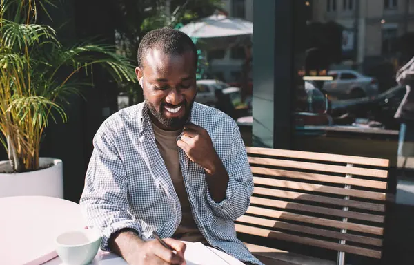 Bluetooth kulaklıklı neşeli erkek öğrenci, eğitim kitabı dinlerken ödev testi yapıyor, kulaklıklı mutlu koyu tenli hippi adam ders kitabında organizasyon planı yazıyor.