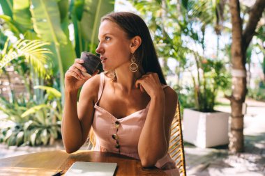 Organizasyon planlaması için ekili eğitim kitabı olan düşünceli bir kadın sokak kafeteryasında kafein içeceği içiyor, elinde kilden kupa tutan bir kadın düşünceli bir şekilde başka tarafa bakıyor.