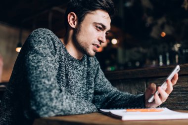 Kafeteryada öğrenim görürken internet yayını okuyan beyaz tenli genç bir adam. Kablosuz sayfaları taramak için akıllı telefon kullanan eğitimli bir blog yazarı.