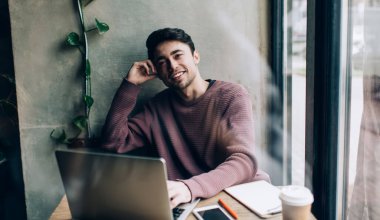 Kafe içinde uzaktan çalışırken kameraya gülümseyen neşeli beyaz bilişim uzmanı portresi. Dijital dizüstü bilgisayarlı mutlu bir erkek blogcu. Kapalı mekanda serbest yaşam tarzının tadını çıkarıyor.