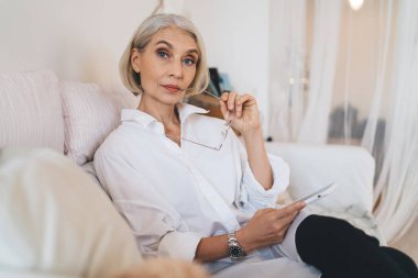 50 yaşında beyaz bir kadın kameraya bakıyor. Kanepede oturan gözlüklü ve akıllı telefonlu modern başarılı kadın. Ev istirahatı ve boş zaman kavramı. Modern geniş dairenin içi bulanık