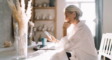 Masada aletlerle çalışan 50 yaşındaki beyaz kadın girişimcinin yan görüntüsü. Küçük işletmeler ve girişimcilik. Modern başarılı kadın. Rafları seramik ürünlerle dolu ev sanatları stüdyosu.