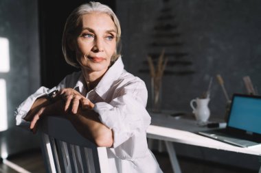50 yaşındaki kadın girişimci sandalyeye oturup gözlerini kaçırıyor. Küçük işletme ve girişimcilik kavramı. Modern başarılı kadın. Karanlık ve aydınlık ev sanatları stüdyosunun içinde.
