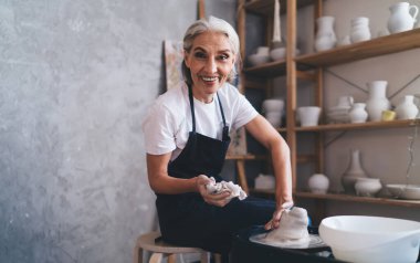 Gülümseyen Kafkas kıdemli kadın heykeltıraş çömlek çarkında kil çömlek yapıyor. Küçük işletme ve girişimcilik kavramı. Ev hobisi, eğlence ve eğlence. Kameraya bakan kadın. Sanat stüdyosu