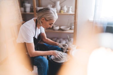 Odaklanmış olgun Avrupalı kadın usta sanat stüdyosundaki çömlek çarkında kil kaplama yapıyor. Küçük işletme ve girişimcilik kavramı. Ev hobisi, eğlence ve eğlence. İş önlüğü giyen kadın.