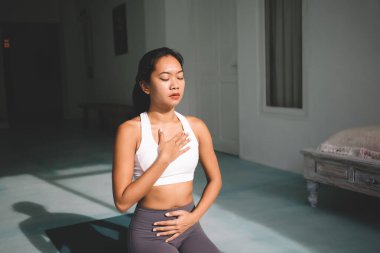 Diyafragmatik nefesle yoga yapan Asyalı kadın geniş evde poz veriyor. Uyum ve akıl sağlığı kavramı. Genç, sakin, güzel, gözleri kapalı, spor kıyafetlerini spor minderinde giyen bir kız.