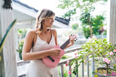 Kafkasyalı genç bir kadın otelin balkonunda ukulele çalıyor. Eğlence, eğlence ve hobi anlayışı. Tatil ve hafta sonu fikri. Dövmeleri olan oldukça konsantre bir kız. Güneşli bir gün. Bali adası