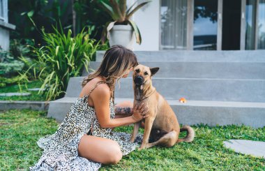 Otelin bahçesinde genç bir kadın sevimli bir köpeği okşuyor. Turizm, tatil ve hafta sonu. Sahibi ve evcil hayvan ilişkisi ve dostluğu. Elbise giyen güzel bir kız. Huzurlu ve huzurlu bir yaşam tarzı. Bali adası