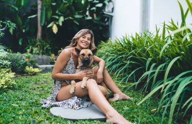 Otelin bahçesinde sevimli köpeğe sarılan neşeli genç bir kadın. Turizm, tatil ve hafta sonu. Sahibi ve evcil hayvan ilişkisi ve dostluğu. Beyaz kız kameraya bakıyor. Idyllic ve sakin yaşam tarzı