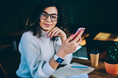 Görme düzeltmesi için optik gözlüklü zeki bir kadının portresi kameraya bakarken bilgilendirici araştırmalar için hücresel uygulama kullanıyor, ders kitabı ve akıllı telefon pozu veren bin yıllık öğrenci.