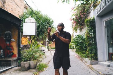 Koyu tenli erkek turist web günlüğü için seyahat içeriği yaratmak için selfie çekerken modern mobil teknolojinin ön kamerasını kullanıyor.