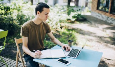 Sıradan kıyafetlerle sokakta ders kitabıyla oturan ve dizüstü bilgisayarla yeni bir proje üzerinde çalışan odaklanmış bir erkek uzaktan kumandalı işçi.