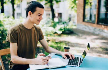 İş sırasında dizüstü bilgisayar kullanırken masada elinde akıllı telefon ve defterle oturan serbest çalışan mutlu bir erkek görülüyor.