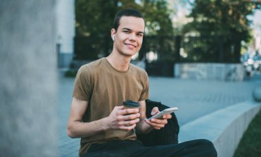 Neşeli erkek, kablosuz kulaklıkla müzik dinliyor ve kameraya bakıp arka planda bir bardak sıcak içecekle taş sınırda otururken akıllı telefonuyla sörf yapıyor.