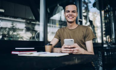 Günlük kıyafetli, elinde not defterleri ve bir fincan kahve ile masada oturan ve gün ışığında kafenin terasında akıllı telefon kullanan pozitif bir adam.