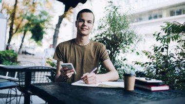 İçerik erkek akıllı telefona göz atıyor ve kameraya bakıyor. Masadaki not defterine notlar alıyor. Kafeteryanın terasındaki bulanık arka plandaki kahveyle birlikte.