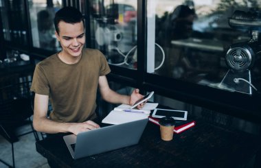 Dizüstü bilgisayarla proje üzerinde çalışan gülümseyen öğrenci ve gündüz vakti kafenin terasında otururken akıllı telefon kullanıyor.