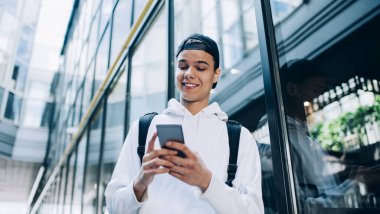 Beyaz kapüşonlu ve sırtında sırt çantasıyla gökdelen yakınlarında yürürken cep telefonuyla internette gezinen neşeli genç adamın alçak açısı.